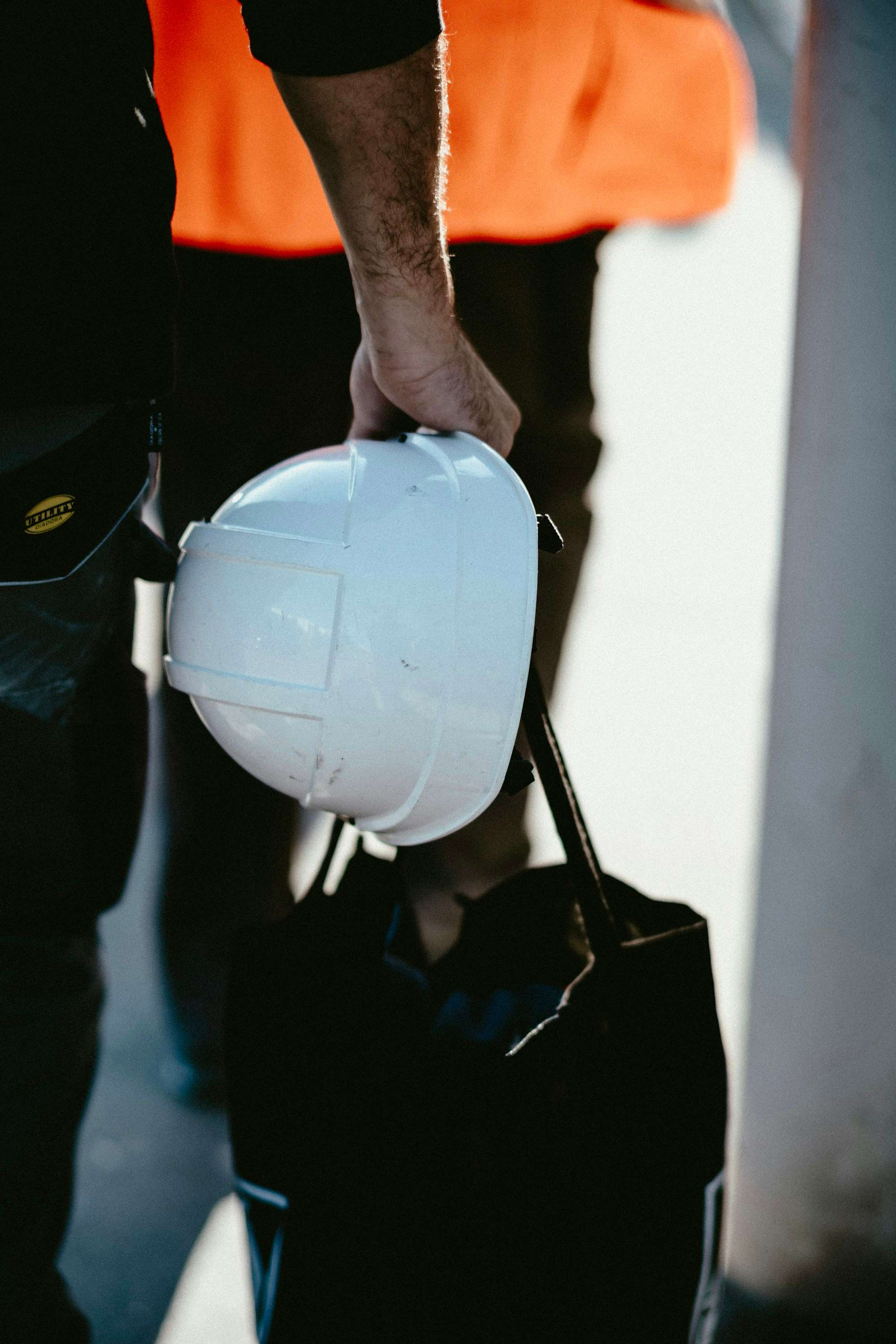 A worker in orange hi-vis carrying a white hard hat and a work bag